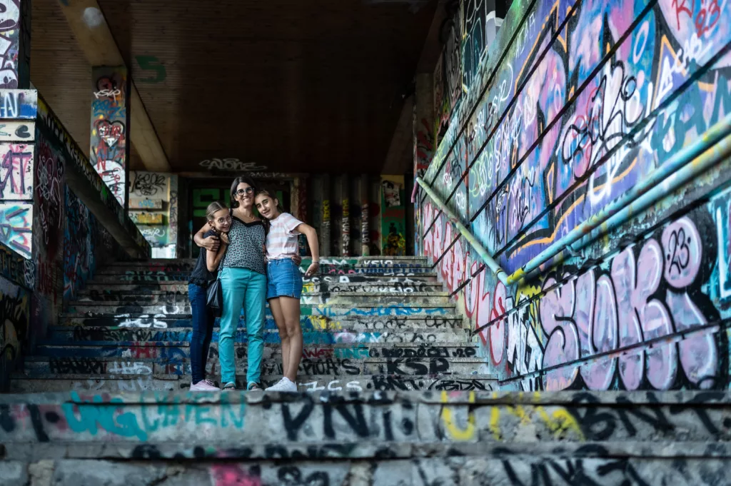 Photo d'une maman enlaçant ses 2 filles adolescentes dans des escaliers plein de graphitis par Simone Photographie